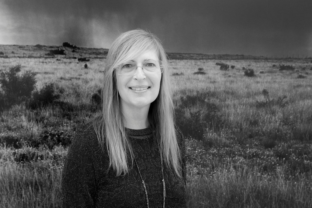Photographie en noir et blanc, en plan semi-rapproché, de Geneviève Cadieux se tenant dos à une image immense représentant une prairie. Portant des lunettes, elle sourit à la caméra. (Afficher en plein écran)