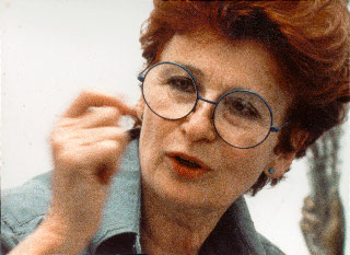 Photographie couleur en plan rapproché de Betty Goodwin, les cheveux roux coupés courts et la main droite levée. Elle porte une chemise en jeans à col ouvert et des lunettes rondes. (Afficher en plein écran)