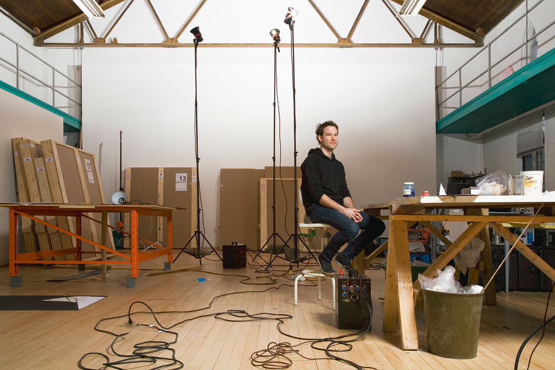 Photographie couleur de Pascal Grandmaison dans un studio d’artiste. Il porte les cheveux courts, un gilet à capuchon noir et un jeans bleu. Assis sur un tabouret à une grande table de travail, il fixe l’objectif et esquisse un sourire. (Afficher en plein écran)