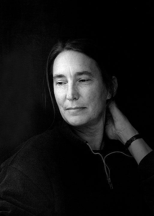 Photographie en noir et blanc à haut contraste sur fond noir de Jenny Holzer portant un gilet foncé. Elle a le regard baissé et sourit discrètement. (Afficher en plein écran)