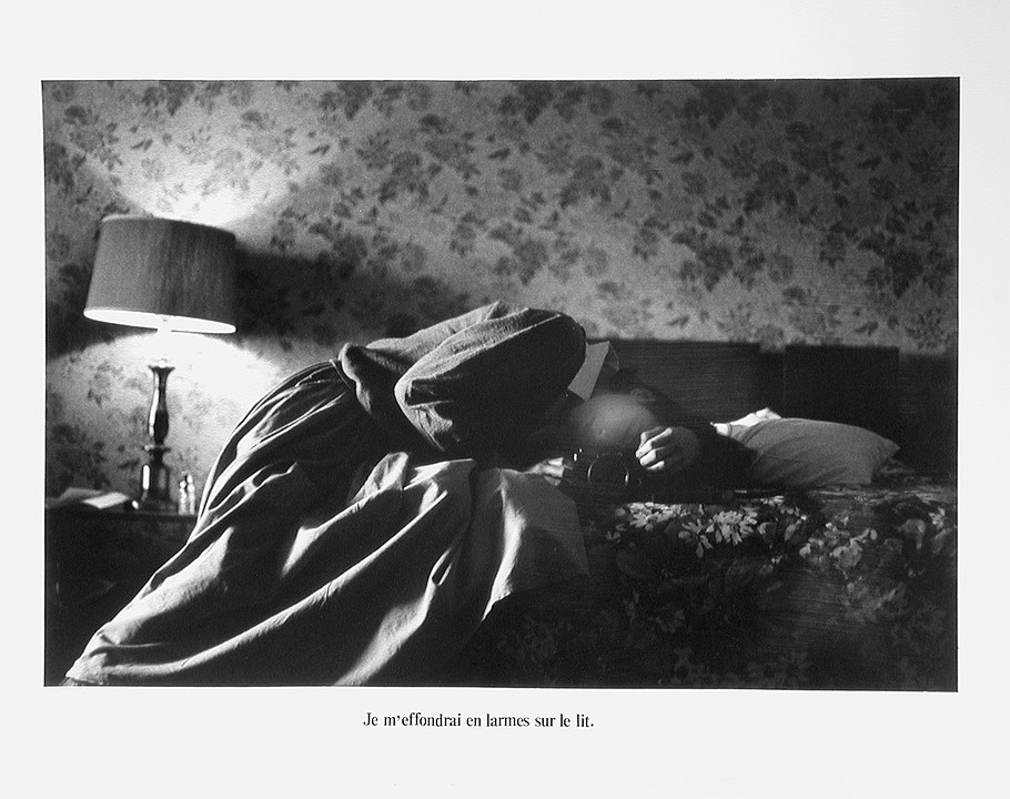 Photographie en noir et blanc d’une jeune femme à moitié étendue sur un lit, sa tête dans le creux de son épaule et tenant un appareil photo. (Afficher en plein écran)