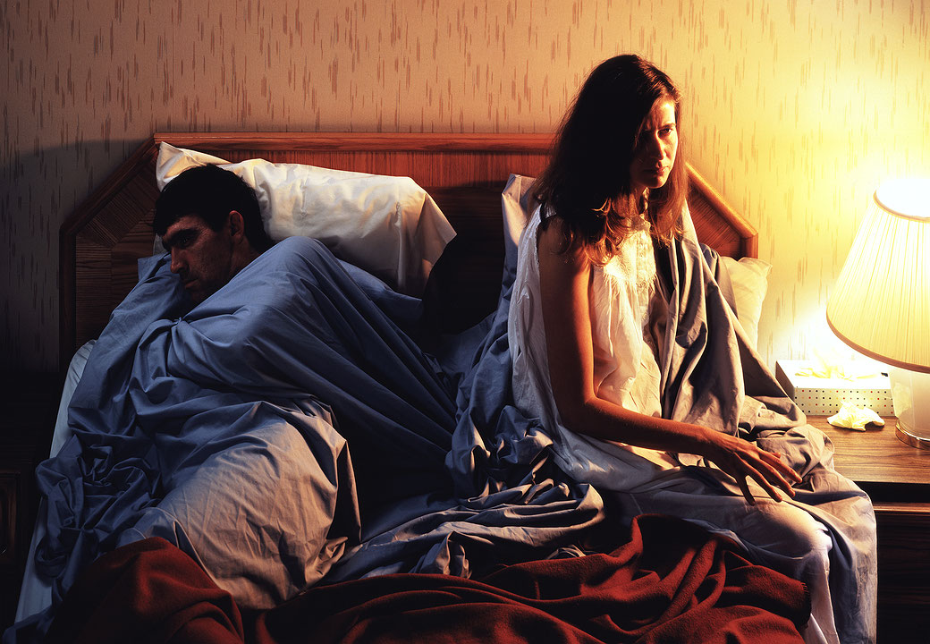 Photographie couleur d’un homme et d’une femme dans une chambre. L’homme est blotti sur le lit, enveloppé d’un drap, le dos tourné. La femme est assise au bord du lit. Elle regarde devant elle, impassible. (Afficher en plein écran)