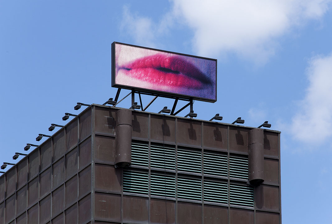 Photographie couleur en plan rapproché d’un panneau lumineux représentant des lèvres féminines, posé sur le toit du Musée d’art contemporain de Montréal, sur un fond de ciel bleu. (Afficher en plein écran)