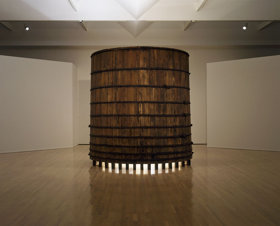 Photographie couleur d’une salle d’exposition dans laquelle une structure cylindrique volumineuse en bois projette une lumière qui émane de sa base. (Afficher en plein écran)