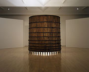 Photographie couleur d’une salle d’exposition dans laquelle une structure cylindrique volumineuse en bois projette une lumière qui émane de sa base.