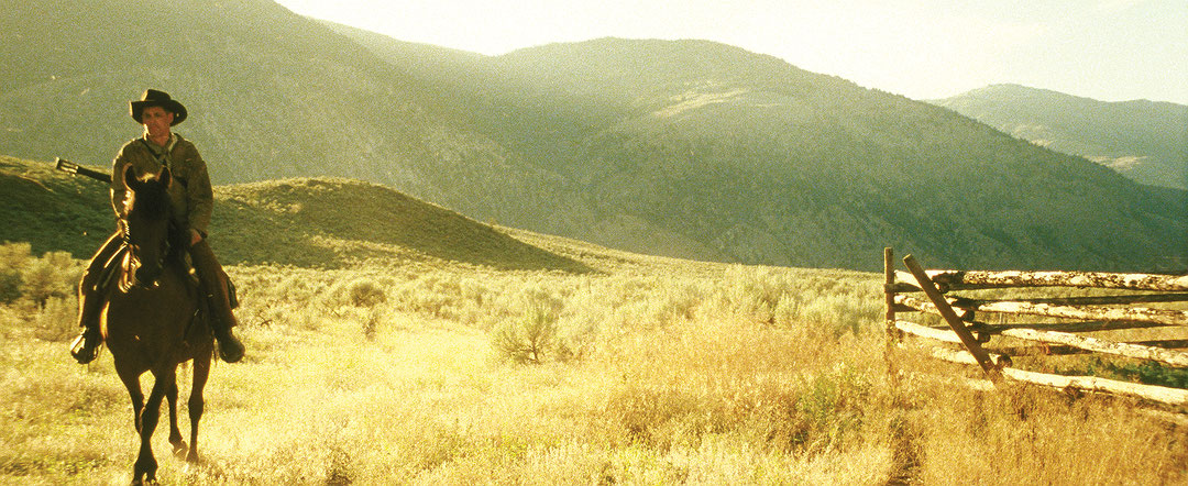 Image couleur. Au premier plan, à gauche, un cow-boy à cheval s’avance dans un vaste champ doré avec sa guitare en bandoulière. Une clôture en bois est située à droite de l’image. Le paysage comporte des montagnes en arrière-plan. (Afficher en plein écran)