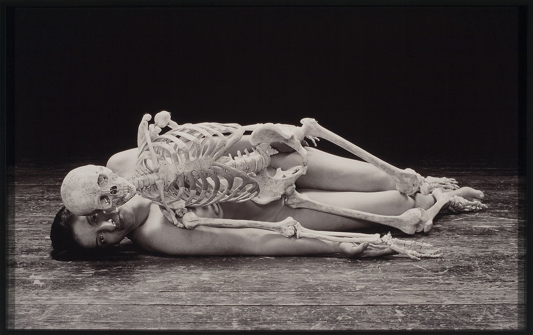 Photographie en noir et blanc de Marina Abramovic nue, couchée sur le côté sur un plancher. Un squelette humain repose sur elle, dans la même position. (Afficher en plein écran)