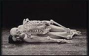 Photographie en noir et blanc de Marina Abramovic nue, couchée sur le côté sur un plancher. Un squelette humain repose sur elle, dans la même position.