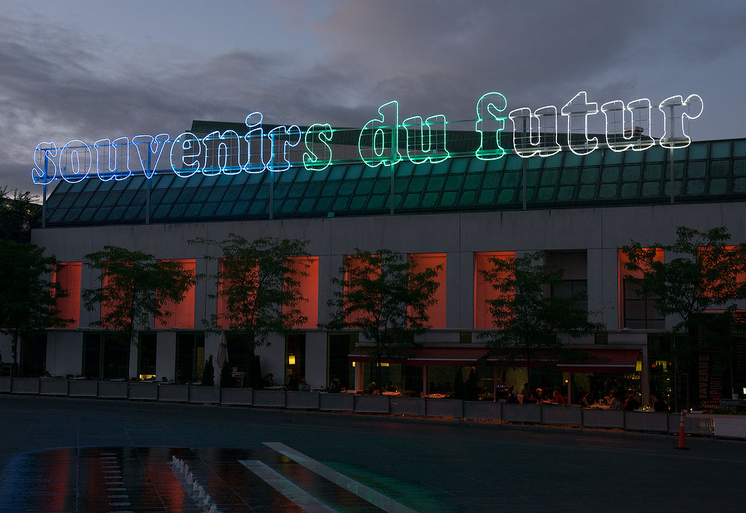 Photographie couleur d’une enseigne au coucher du soleil sur laquelle apparaissent les mots souvenirs du futur en néons colorés au-dessus du Musée d’art contemporain de Montréal. Les parois de l’édifice sont éclairées d’une lumière rouge. Des gens sont assis à une terrasse. (Afficher en plein écran)