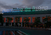 Photographie couleur d’une enseigne au coucher du soleil sur laquelle apparaissent les mots souvenirs du futur en néons colorés au-dessus du Musée d’art contemporain de Montréal. Les parois de l’édifice sont éclairées d’une lumière rouge. Des gens sont assis à une terrasse.