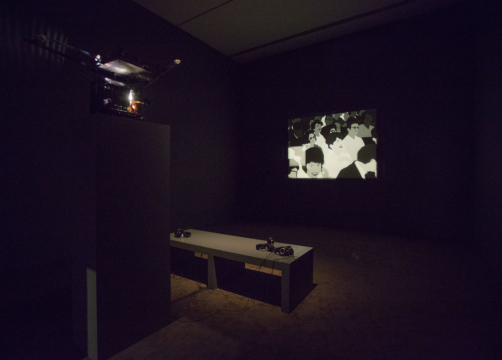 Photographie couleur d’une salle d’exposition sombre, où un projecteur renvoie une image en noir et blanc d’un film d’animation sur un écran au mur. Entre les deux, sur un banc, sont disposés des écouteurs. (Afficher en plein écran)