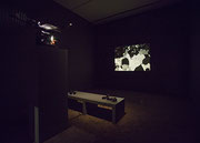 Photographie couleur d’une salle d’exposition sombre, où un projecteur renvoie une image en noir et blanc d’un film d’animation sur un écran au mur. Entre les deux, sur un banc, sont disposés des écouteurs.